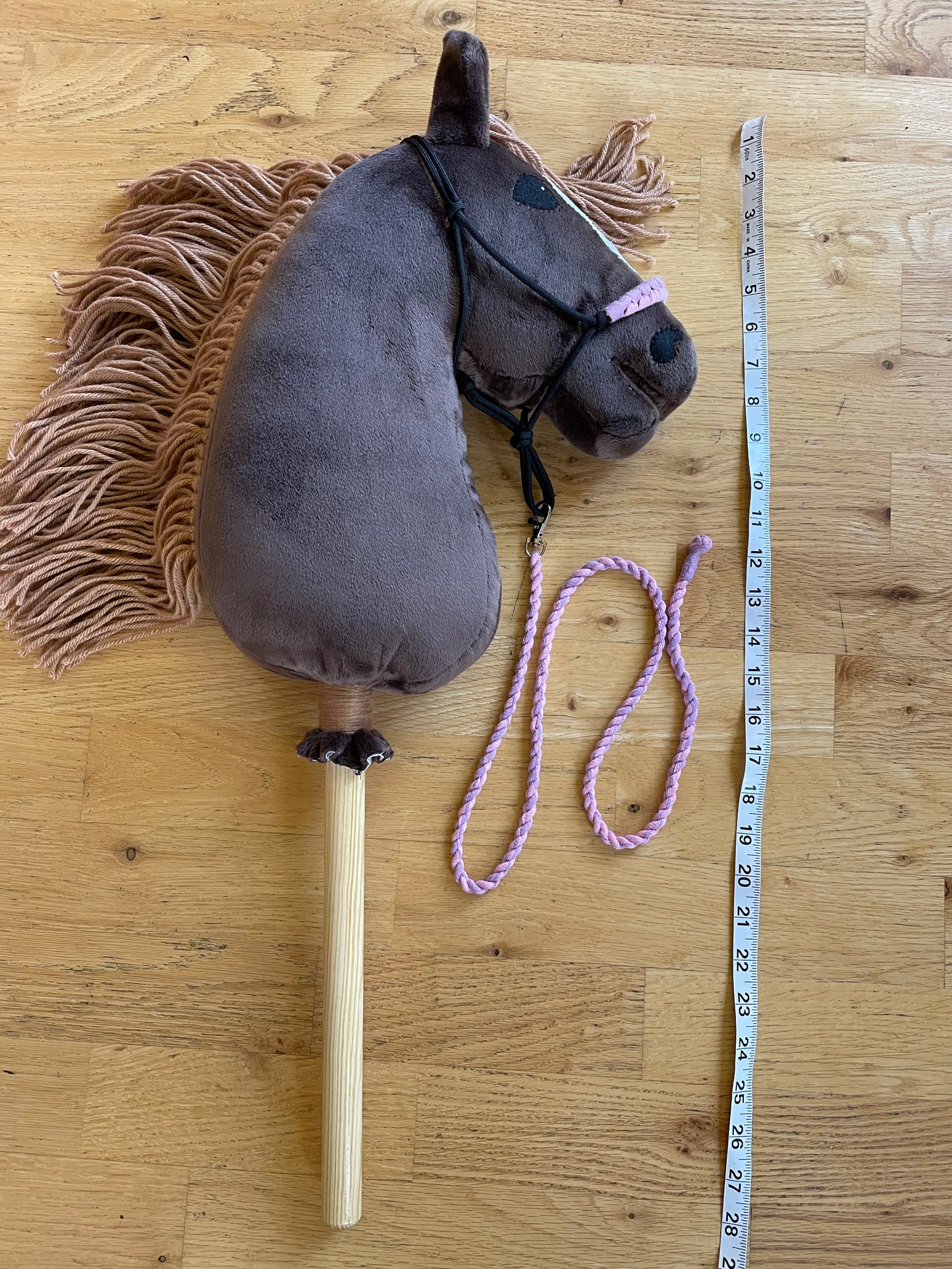 Brown hobby horse plush toy with a measuring tape for scale on a wooden surface.