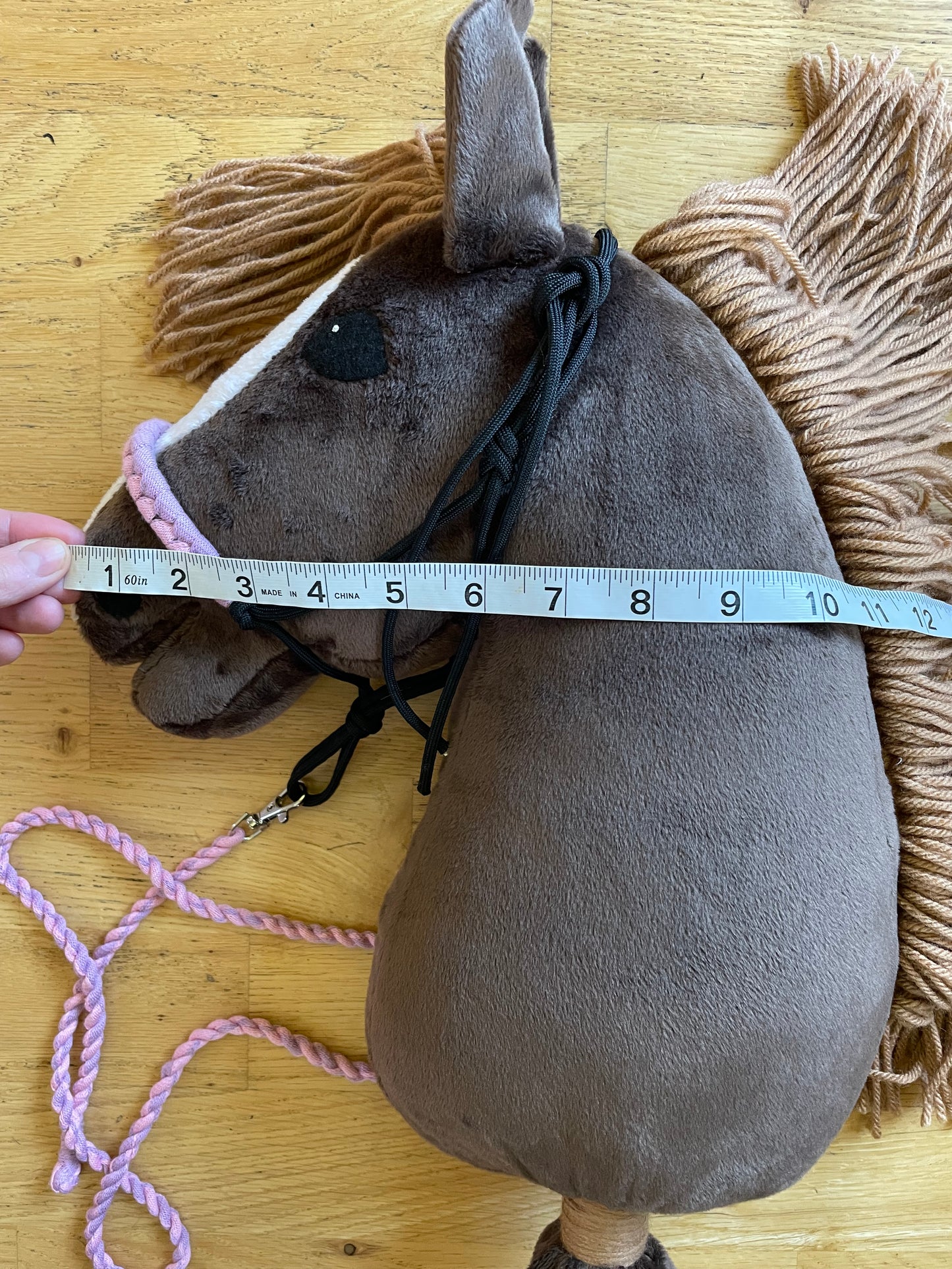 Brown plush hobby horse toy with a measuring tape on a wooden floor. Horse has a rope halter on.