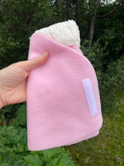 A hand holds a pink fleece hobby horse blanket with a white sherpa lining and a white hook and loop strap, set against a background of green outdoor foliage.