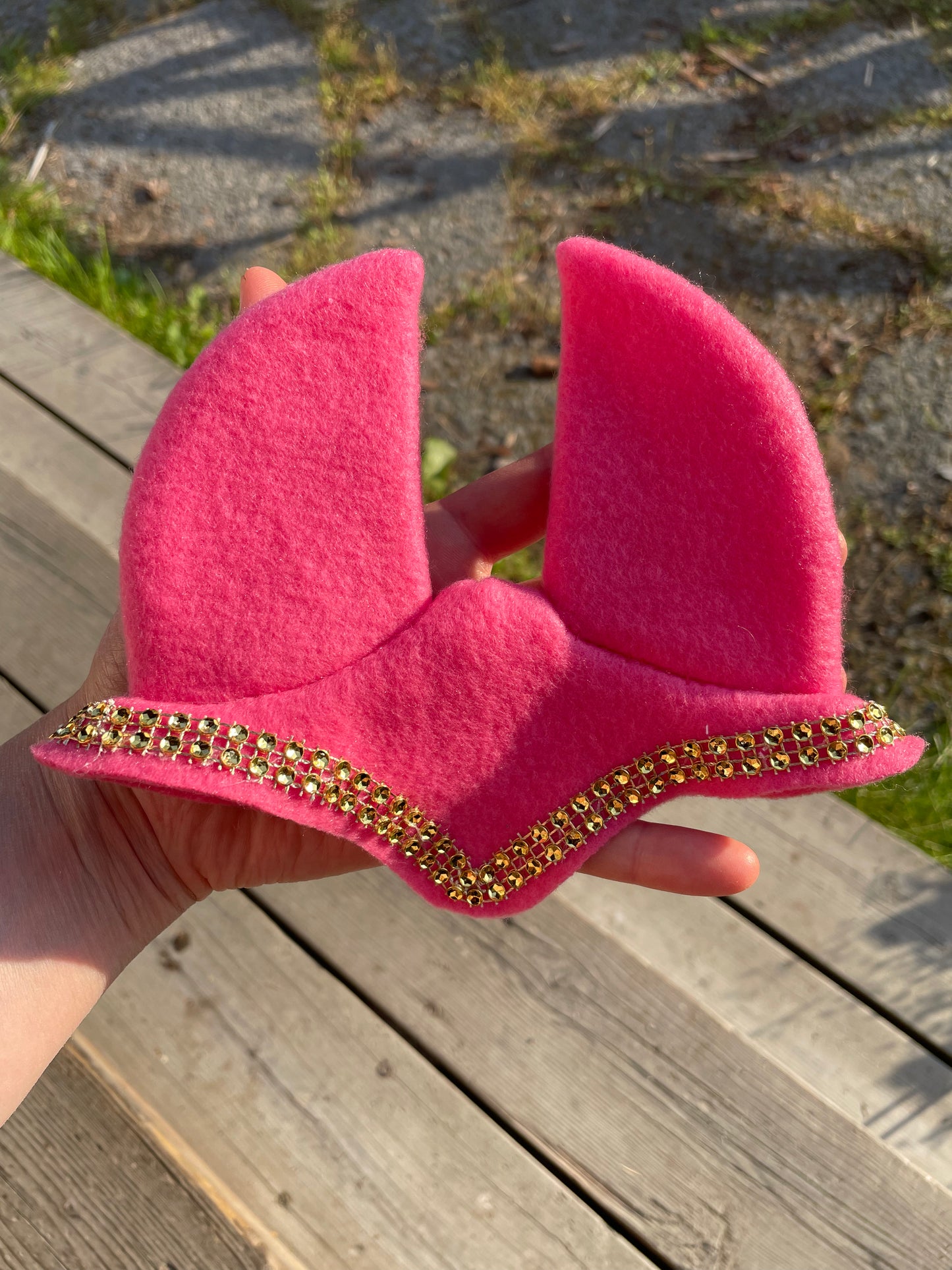 A bright pink felt hobby horse ear bonnet with a decorative gold rhinestone border, held in a hand against a background of wooden planks and gravel.