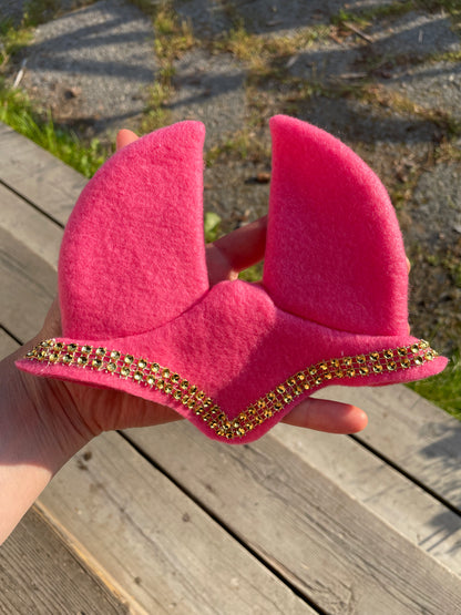 A bright pink felt hobby horse ear bonnet with a decorative gold rhinestone border, held in a hand against a background of wooden planks and gravel.