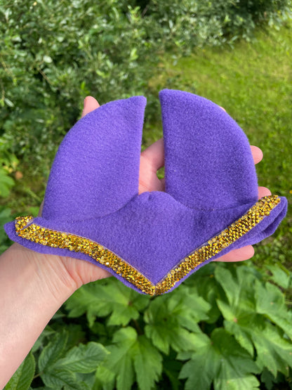 A hand holding a purple fleece hobby horse ear bonnet with a gold glitter trim, set against a background of green garden foliage.