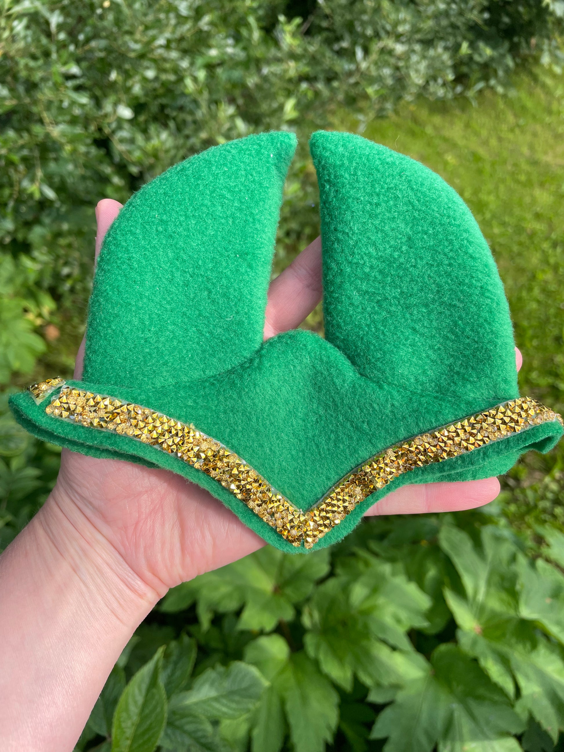 A hand holding a green fleece hobby horse ear bonnet with a gold glitter trim, set against a background of green outdoor foliage.