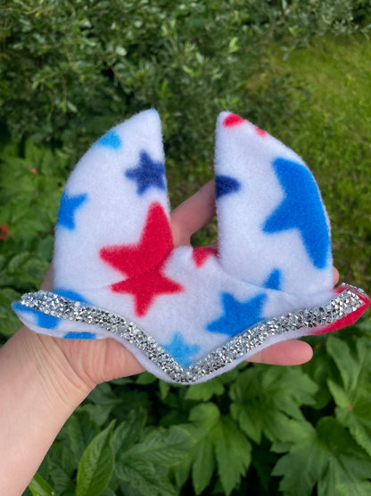 A hand holding a white fleece hobby horse ear bonnet decorated with red, blue, and navy star patterns and a silver glitter trim, set against a background of green garden foliage.