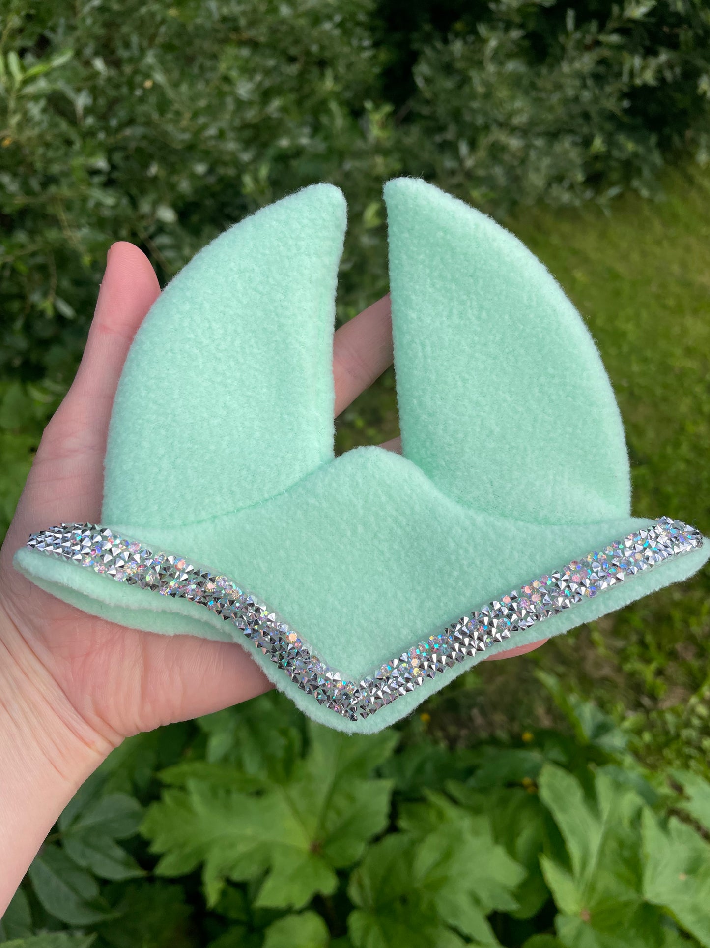 A mint green fleece hobby horse ear bonnet with a silver glitter trim, held in a hand against a background of green garden foliage.