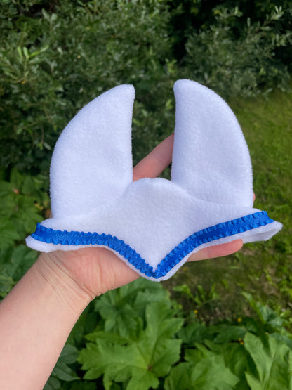 A hand holding a white fleece hobby horse ear bonnet with a blue zigzag trim, set against a background of green garden foliage.