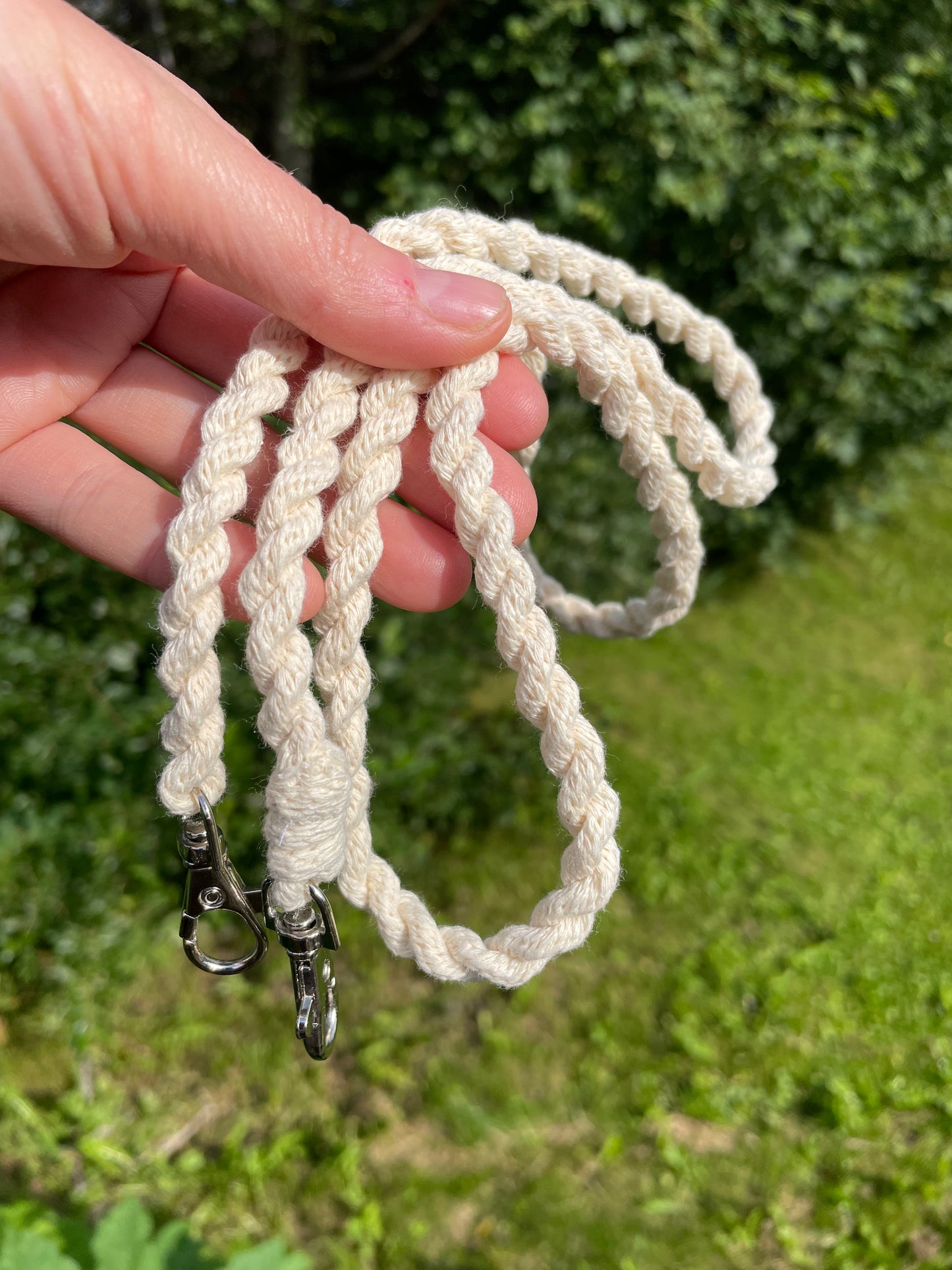 A hand holding a set of off-white braided hobby horse reins with silver metal clips, set against a blurred green outdoor background.