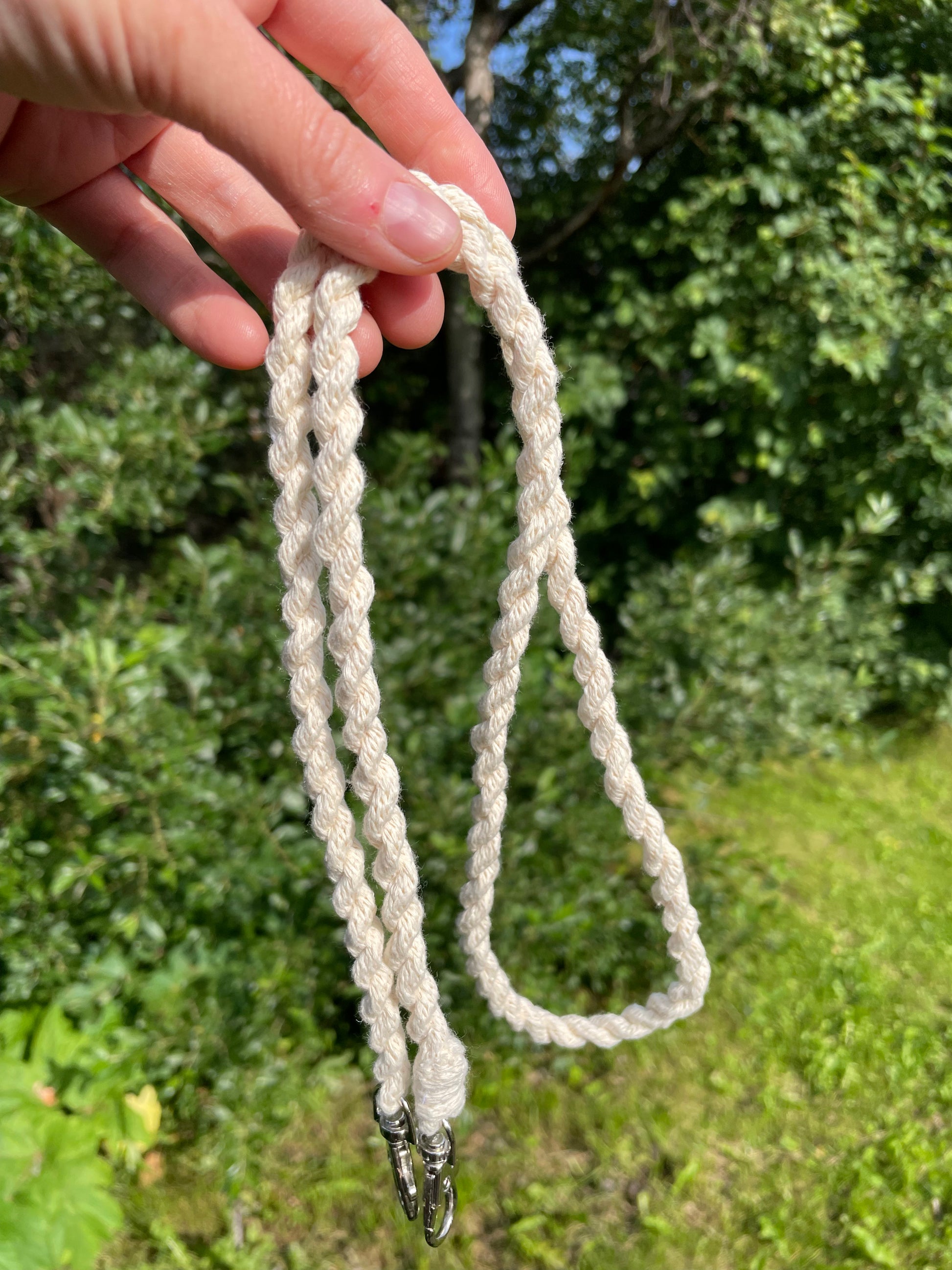A close-up of a hand holding a cream-colored braided hobby horse reins outdoors with a green garden background. The lead rope has silver metal clasps at the end.