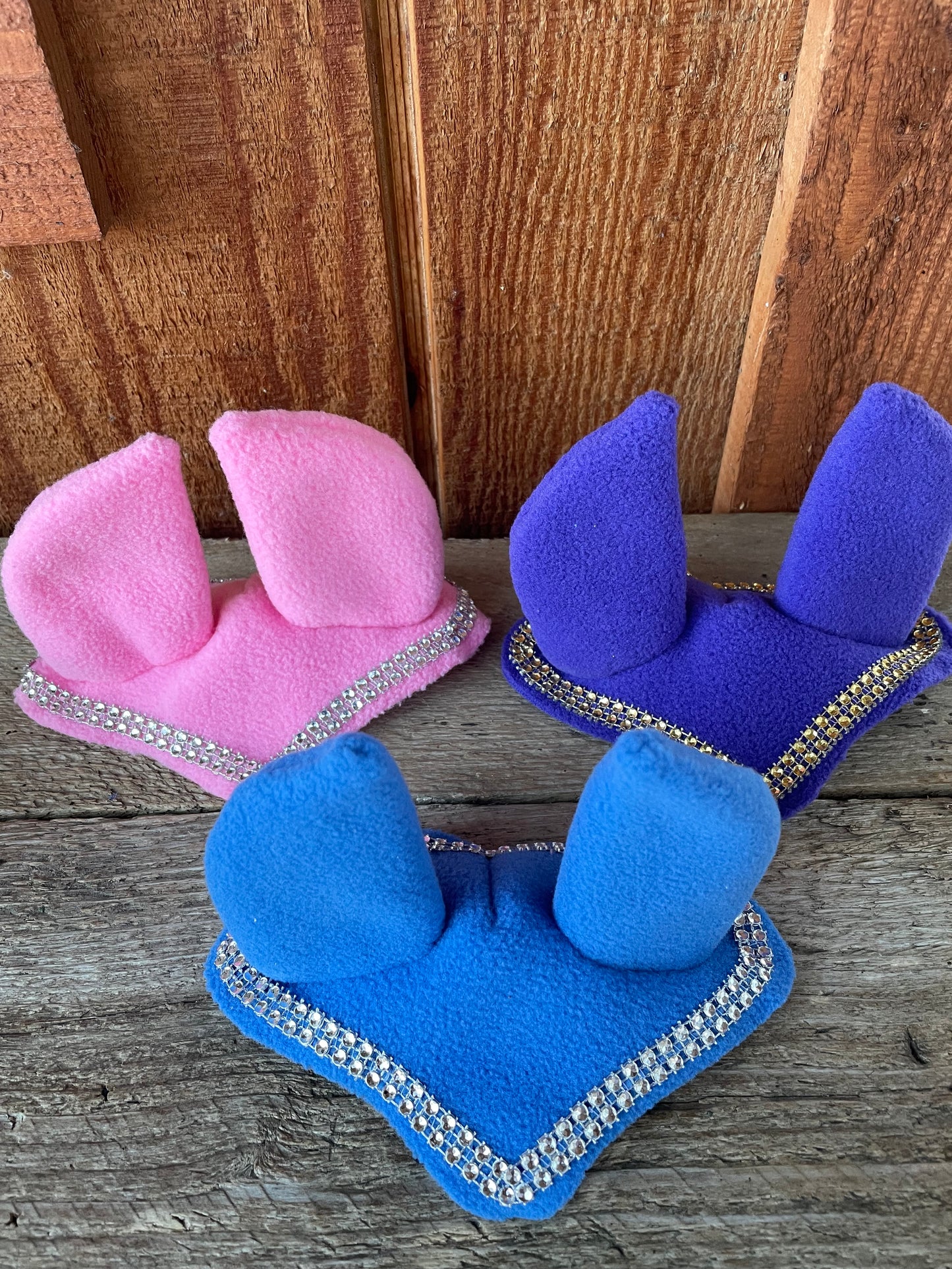 Hobby horse ear bonnets in blue, pink and purple. They have bling on them. Background is wooden wall.