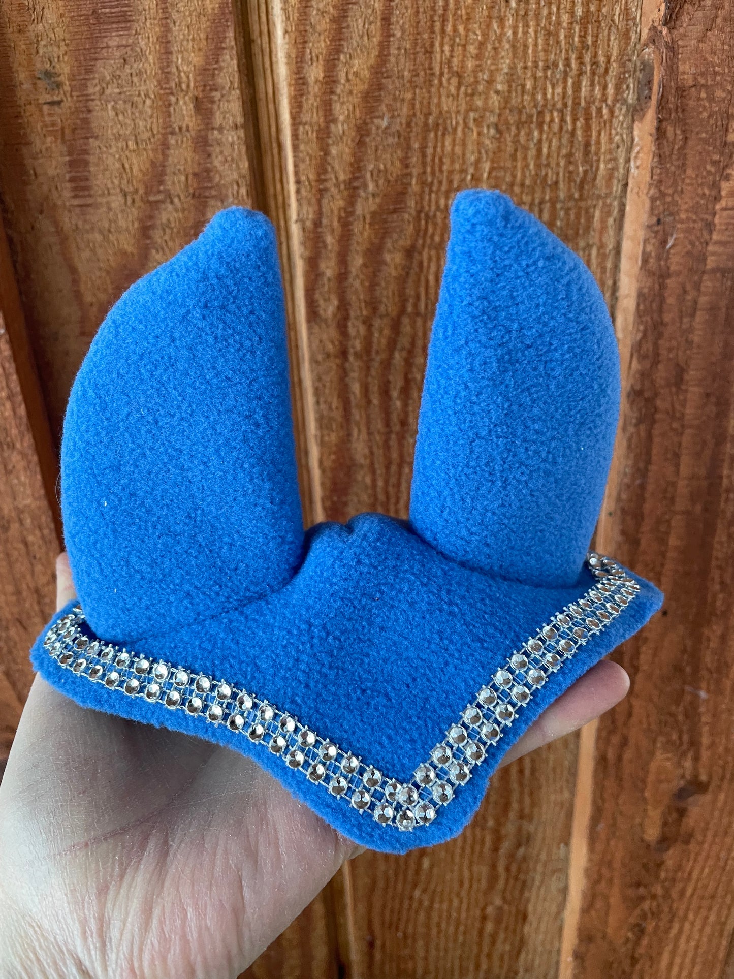 Hobby horse ear bonnet in blue. It has silver bling on it. Background is wooden wall.