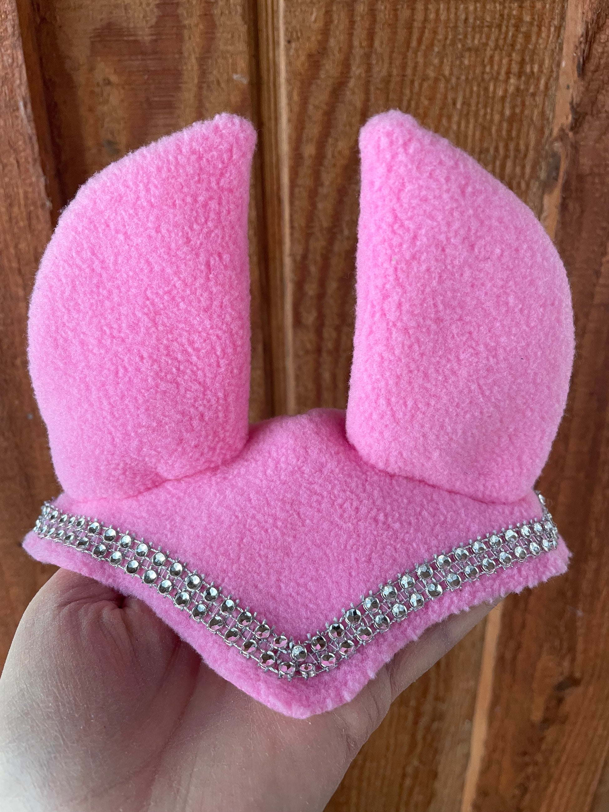 Hobby horse ear bonnet in pink. It has silver bling on it. Background is wooden wall.