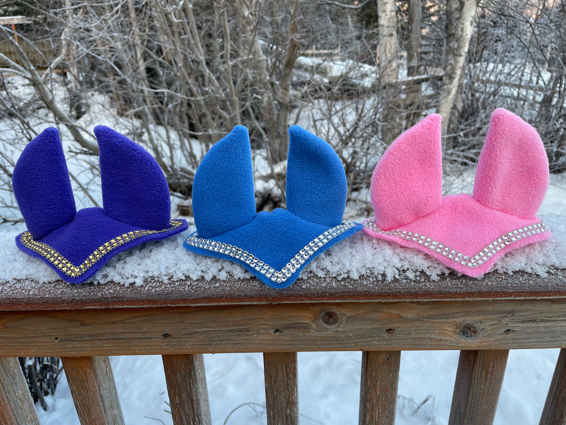 Hobby horse ear bonnet in pink, blue and dark purple. They have bling on them. Background is snowy picture.