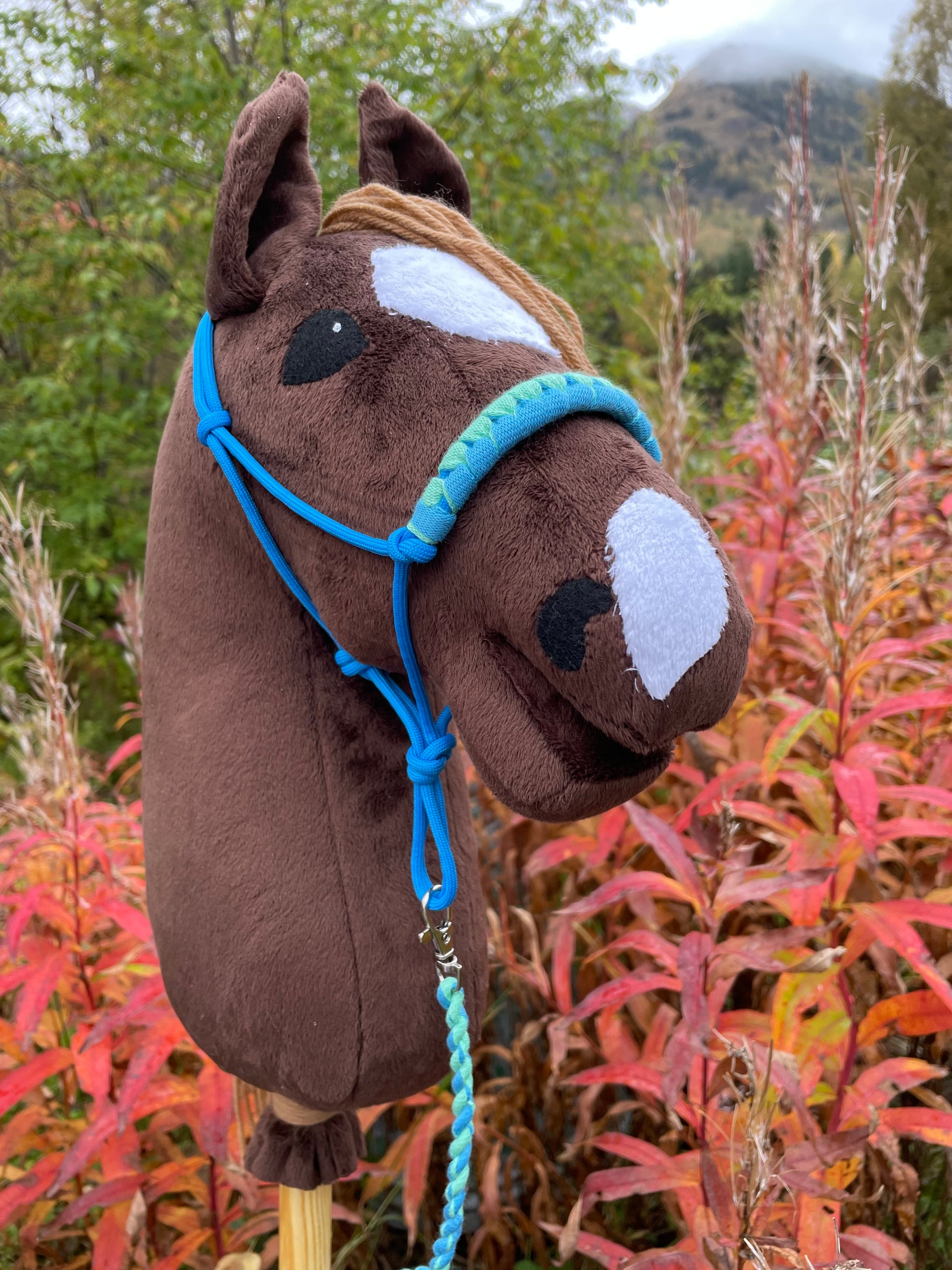 Brownb plush hobby horse toy with a blue halter against a natural background with trees and red foliage.