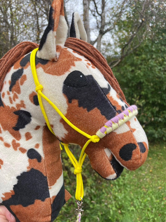 Plush hobby horse pinto toy with a yellow rope halter on against a green outdoor background