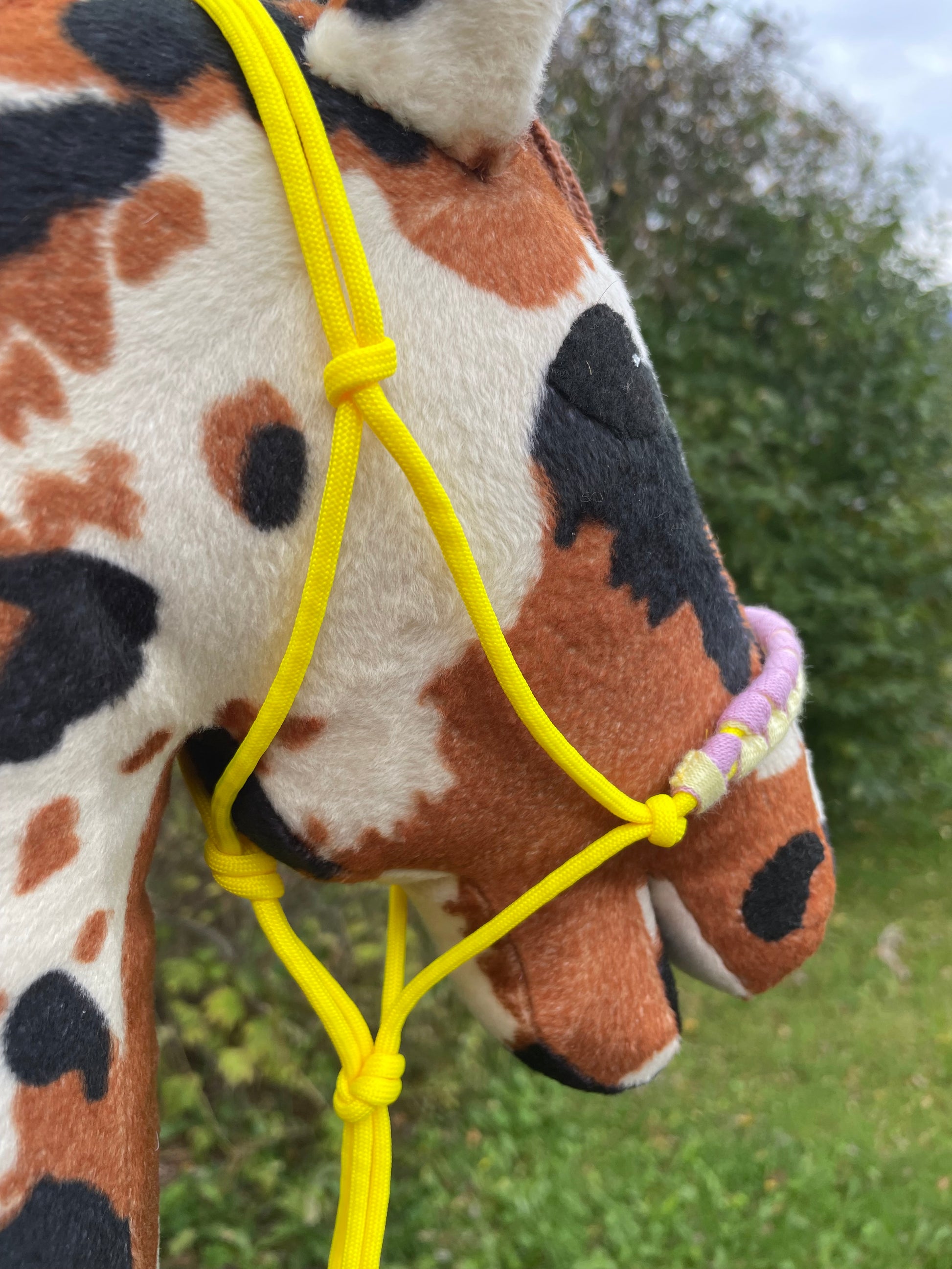 Plush hobby horse toy with a yellow rope halter on against a green outdoor background