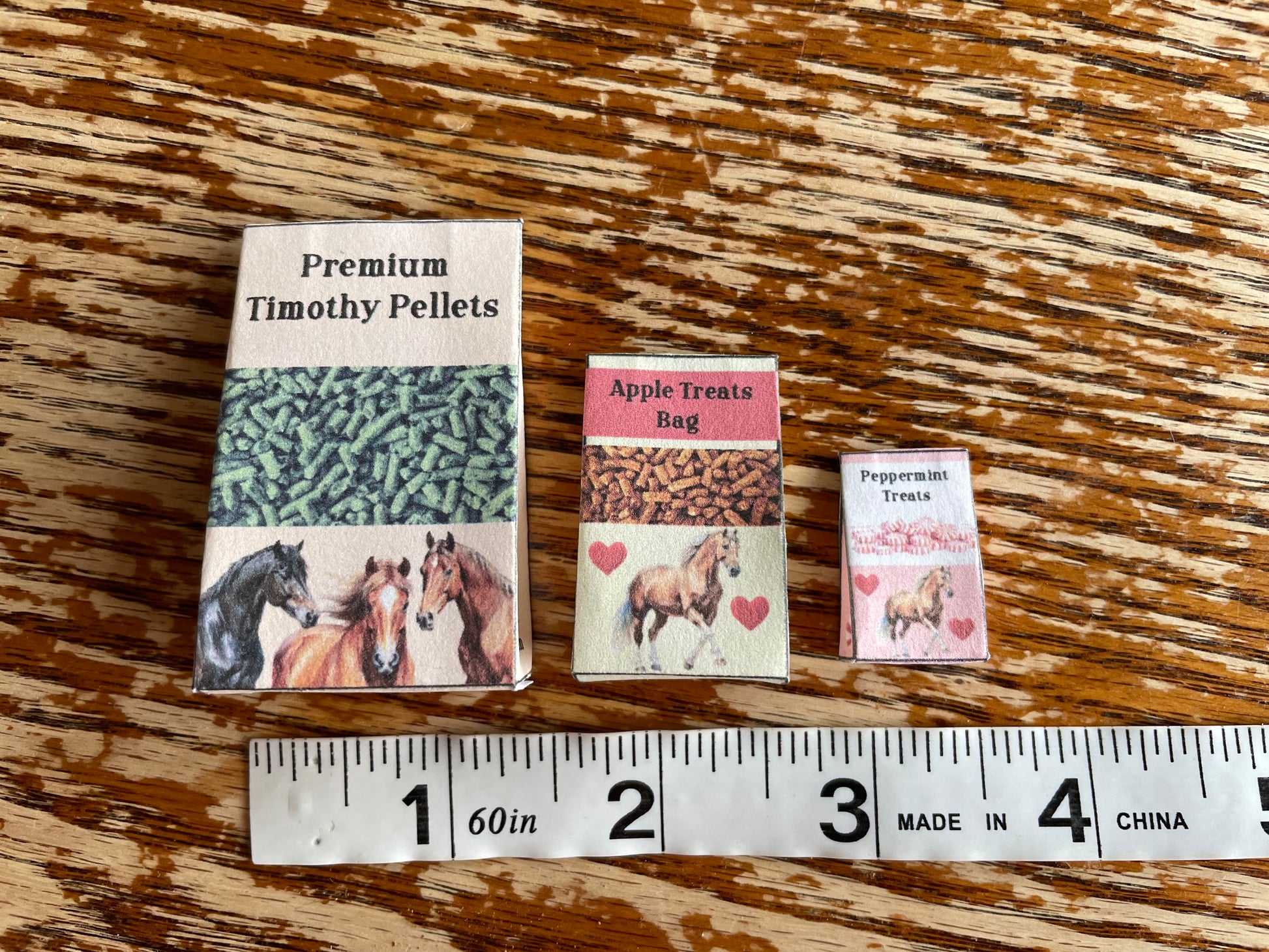 Three miniature packages of horse treats on a wooden surface with a ruler for scale.