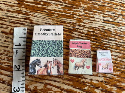 Three small packages labeled 'Premium Timothy Pellets', 'Apple Treats Bag', and 'Peppermints Treats' on a wooden surface with a ruler for scale.